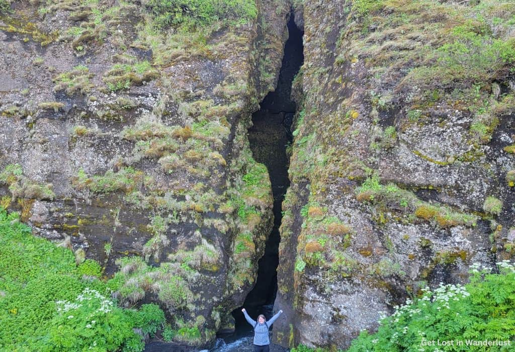 A person standing at the entrance to Gljúfrabúi, which is a small cave opening between two tall cliff edges. The cliff is rocky but surrounded by greenery.