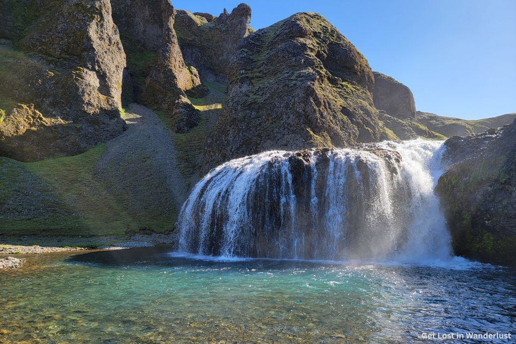The beautiful Stjórnarfoss waterfall, with the water filling into the pool below. The water is a stunning turquoise color which contrasts with the green landscape.