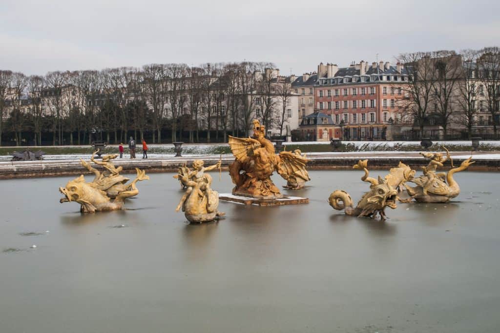 Gold fountain featuring 4 statues and a larger statue in the center. In the background is the town of Versailles and trees that lost their leaves due to winter.