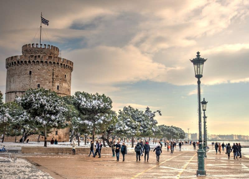 A street in Thessaloniki with snow covered trees and people wearing winter coats. In the background is a tower.