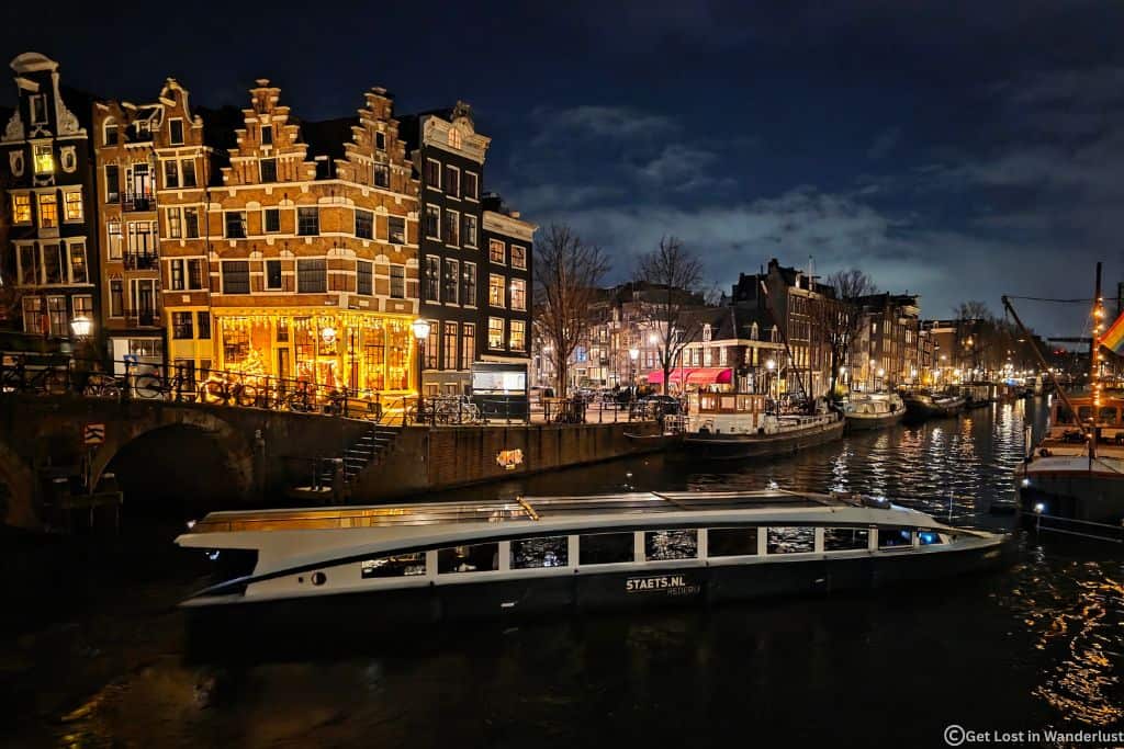 A boat sailing at night through the Amsterdam canals, surrounded by the lit up canal buildings.