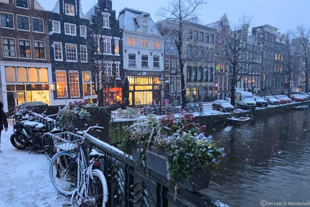 Snowy day during January in Amsterdam. Photo shows the canal with snow falling on a bridge covering a bicycle. In the distance are the historic canal houses.