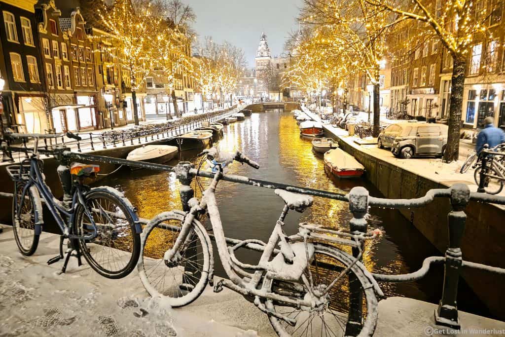 Amsterdam canal with Christmas lights in the trees and snow on the ground.