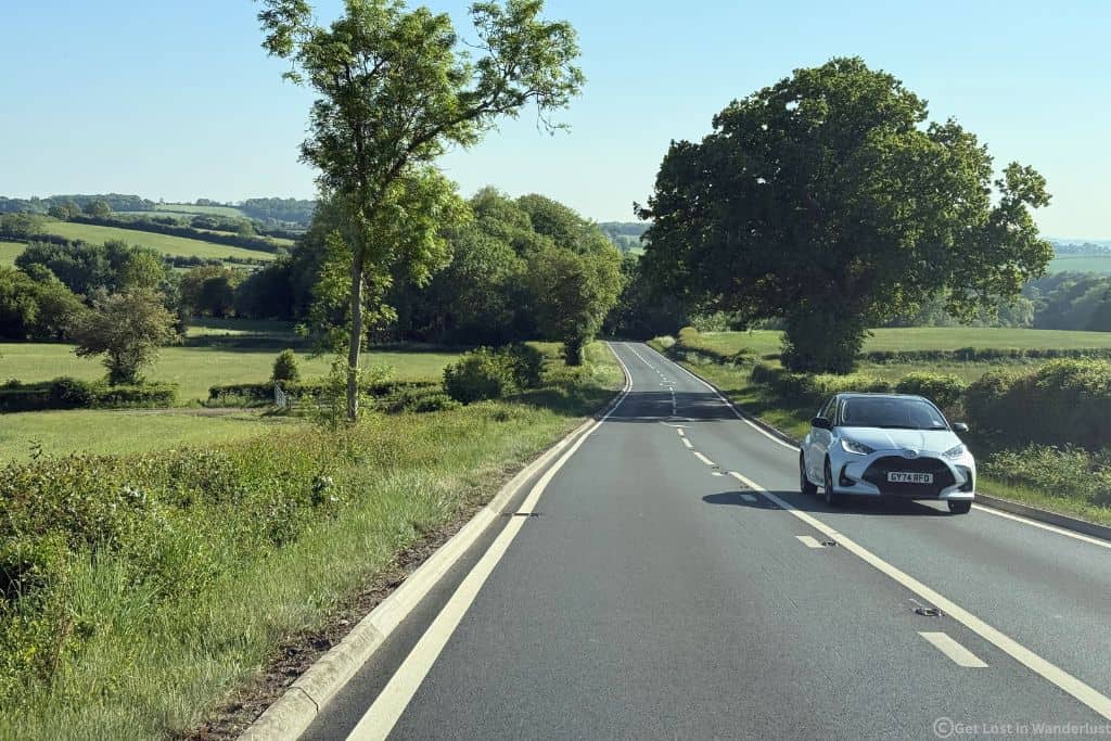A road in England showing a car driving on the left hand side and the scenic views of the English countryside.