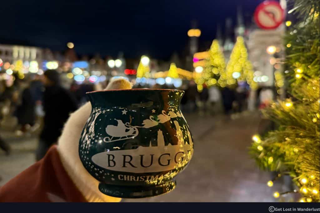 Bruges Christmas Market mug with mulled wine held in front of glowing Christmas lights and festive market stalls at night in Bruges, Belgium.