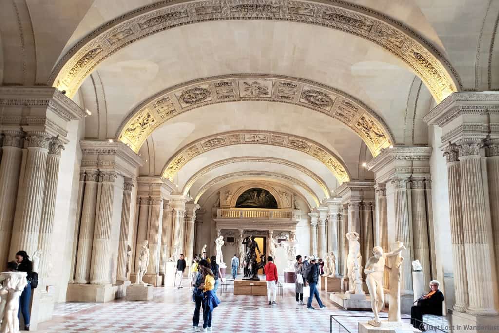 Visitors touring the Louvre Museum’s sculpture galleries beneath vaulted arches in Paris. The Louvre is worth visiting on a trip to Paris, as its one of the best museums in the entire world.