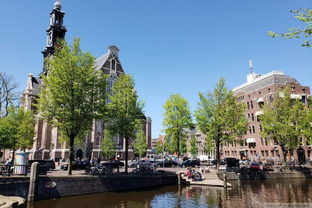 Westerkerk and canal in Amsterdam’s Jordaan neighborhood on a sunny day, with historic buildings, trees, bicycles, and people along the waterfront. This is one of the safest places to stay in Amsterdam.