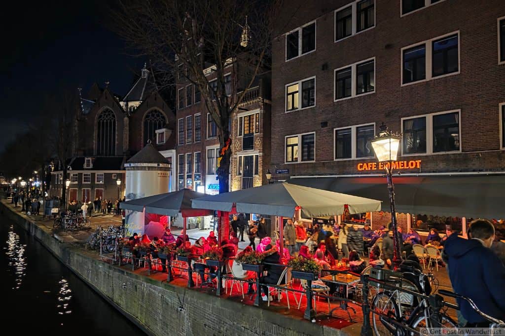 Canal-side café in Amsterdam’s De Wallen Red Light District at night, with outdoor seating, bicycles, and pedestrians.