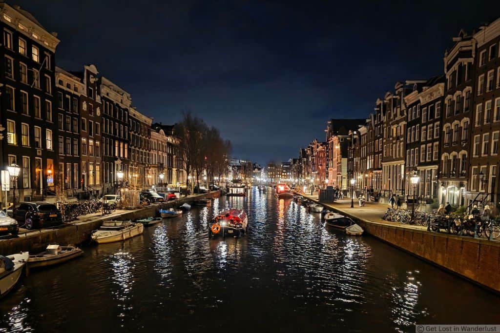 Amsterdam canal at night lined with illuminated historic canal houses, boats, bicycles, and pedestrians.
