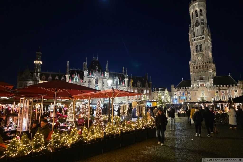 Bruges Christmas Market at night in Markt Square with festive lights, outdoor dining, crowds, and the illuminated Belfry of Bruges.