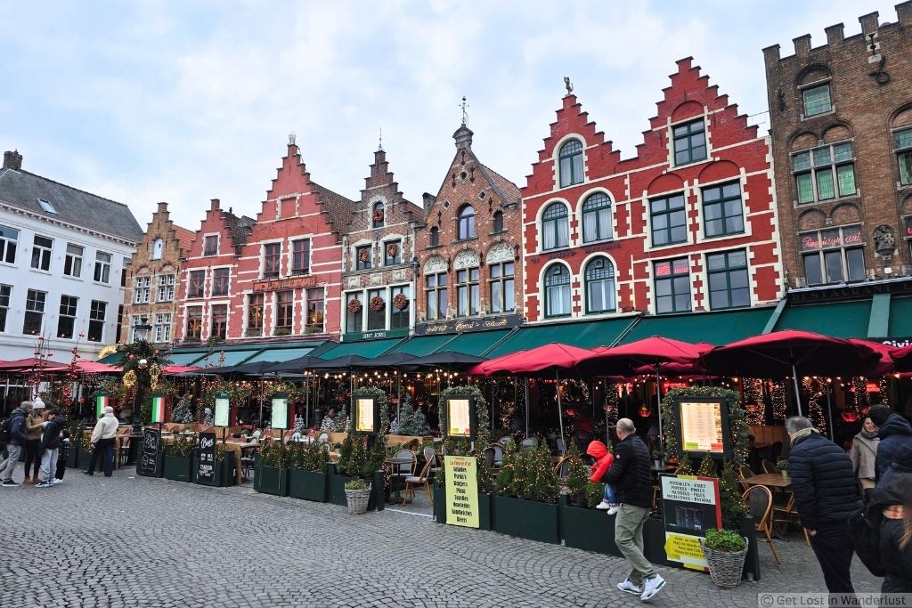 Historic buildings and outdoor cafés decorated for Christmas in Bruges, Belgium, with festive lights and greenery in the city center.
