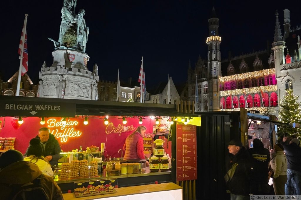 Belgian waffle and crêpe stand at the Bruges Christmas Market at night, with festive lights and historic buildings in Markt Square.