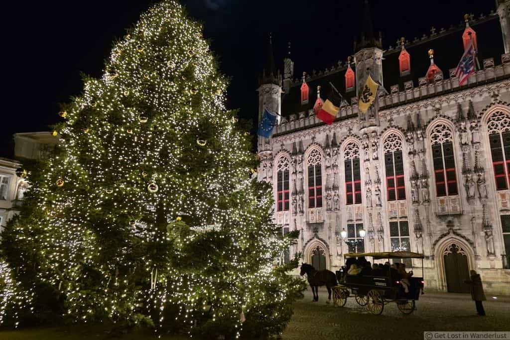Large illuminated Christmas tree beside Bruges City Hall at night, with a horse-drawn carriage in Burg Square, Belgium.