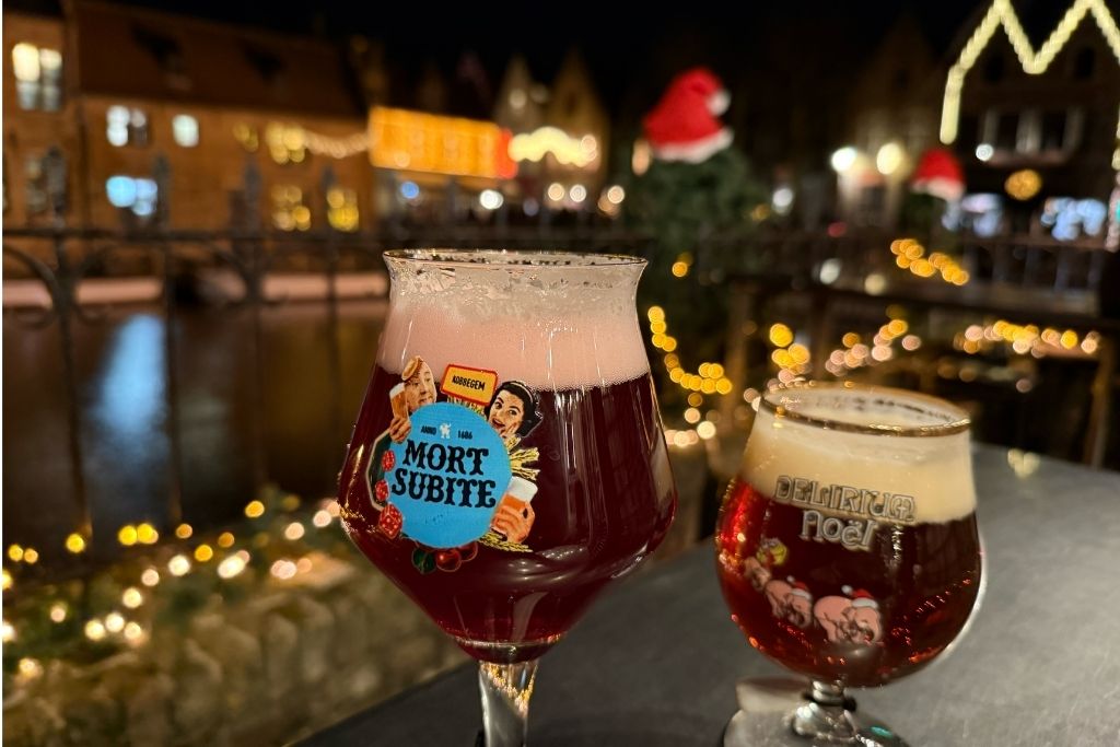 Two festive Belgian beers beside a Bruges canal at night, with Christmas lights and decorated historic buildings in the background.