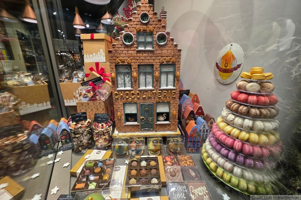 Festive Belgian chocolate shop window in Bruges with boxed chocolates, a chocolate house display, and a colorful tower of macarons.
